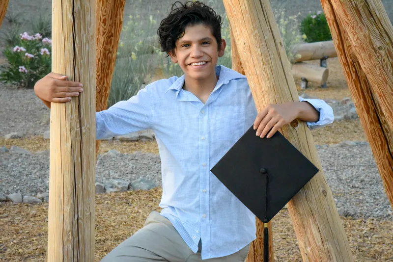 Graduado joven con camisa azul sosteniendo birrete entre postes de madera en una sesión de fotos profesional.