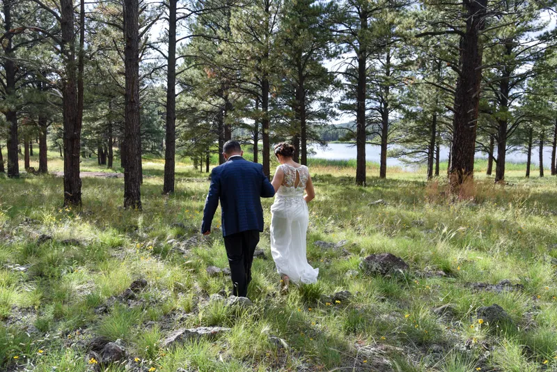 Pareja de novios caminando por un bosque con pinos y un lago al fondo en sesión fotográfica.