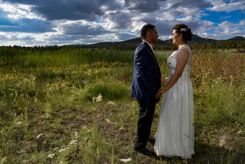 Novios mirándose frente a frente en un campo verde con montañas y nubes dramáticas en el fondo.