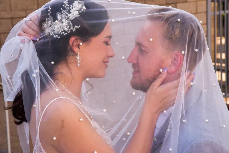 Pareja de novios mirándose fijamente bajo un velo blanco decorado con perlas pequeñas durante su boda.