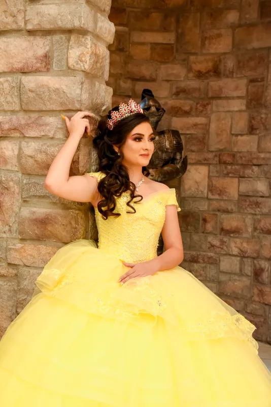 Quinceañera con vestido amarillo y corona posando elegantemente contra una pared de piedra rústica.