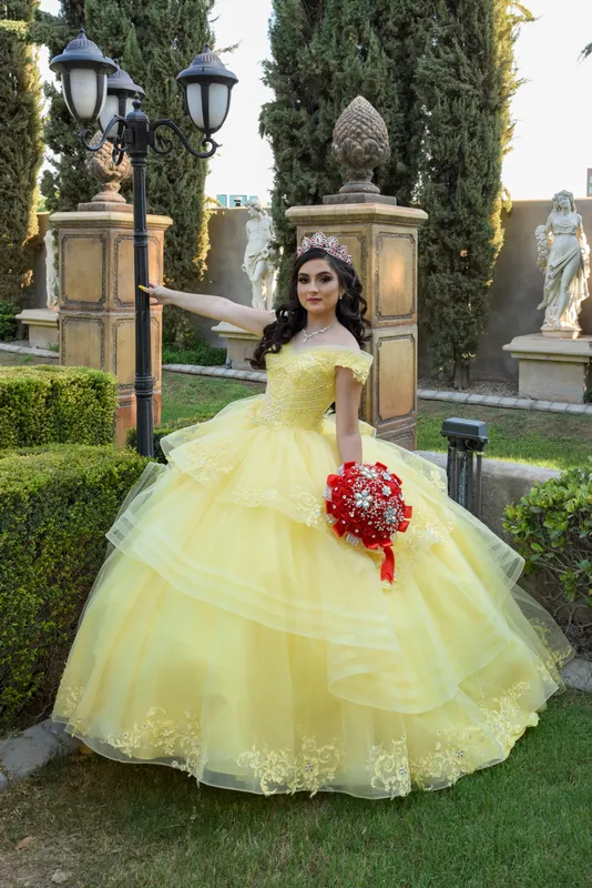 Quinceañera con vestido amarillo y ramo rojo posando junto a una farola antigua en un jardín.