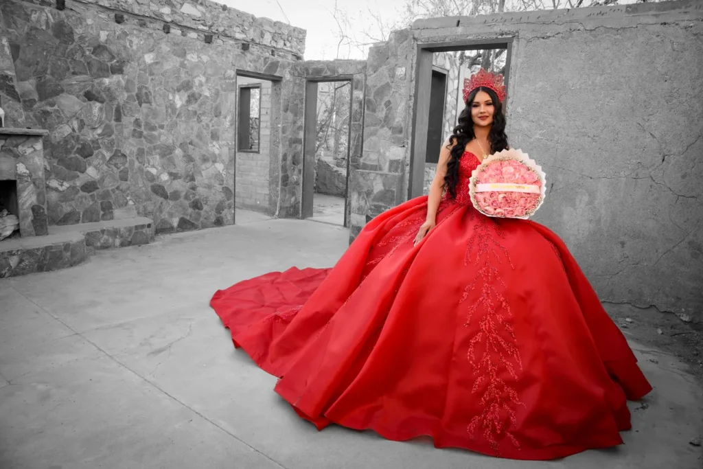 Quinceañera usando vestido de gala rojo brillante y corona, sosteniendo un ramo de rosas frente a ruinas de piedra.